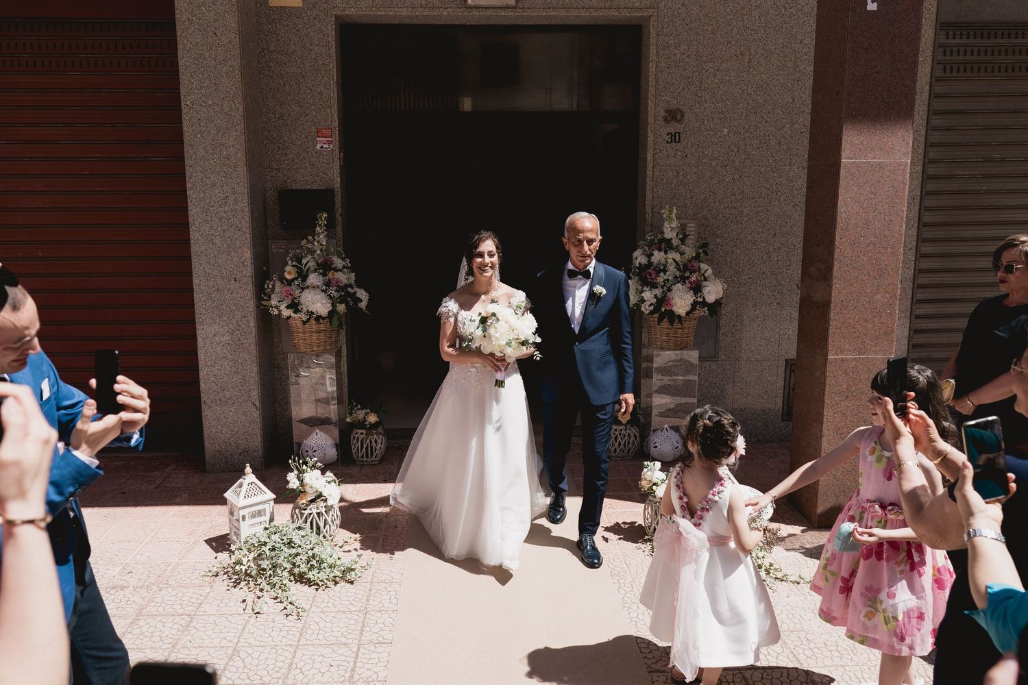 Wedding party gathered outside a building entrance with floral decorations.