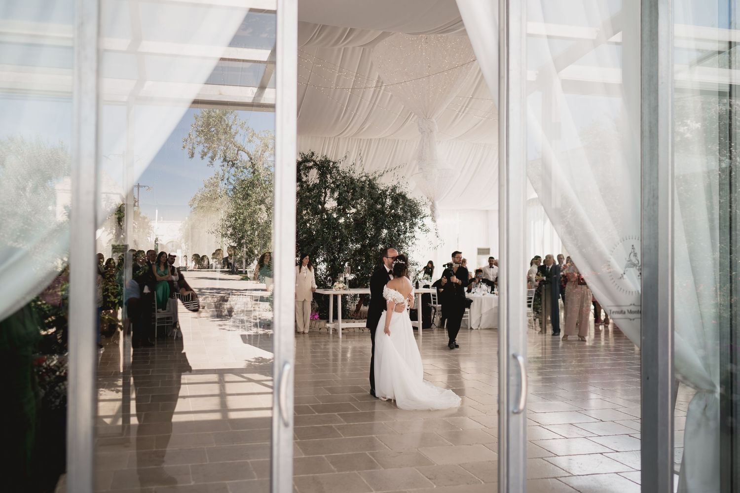 Wedding celebration viewed through glass doors with flowing white curtains.