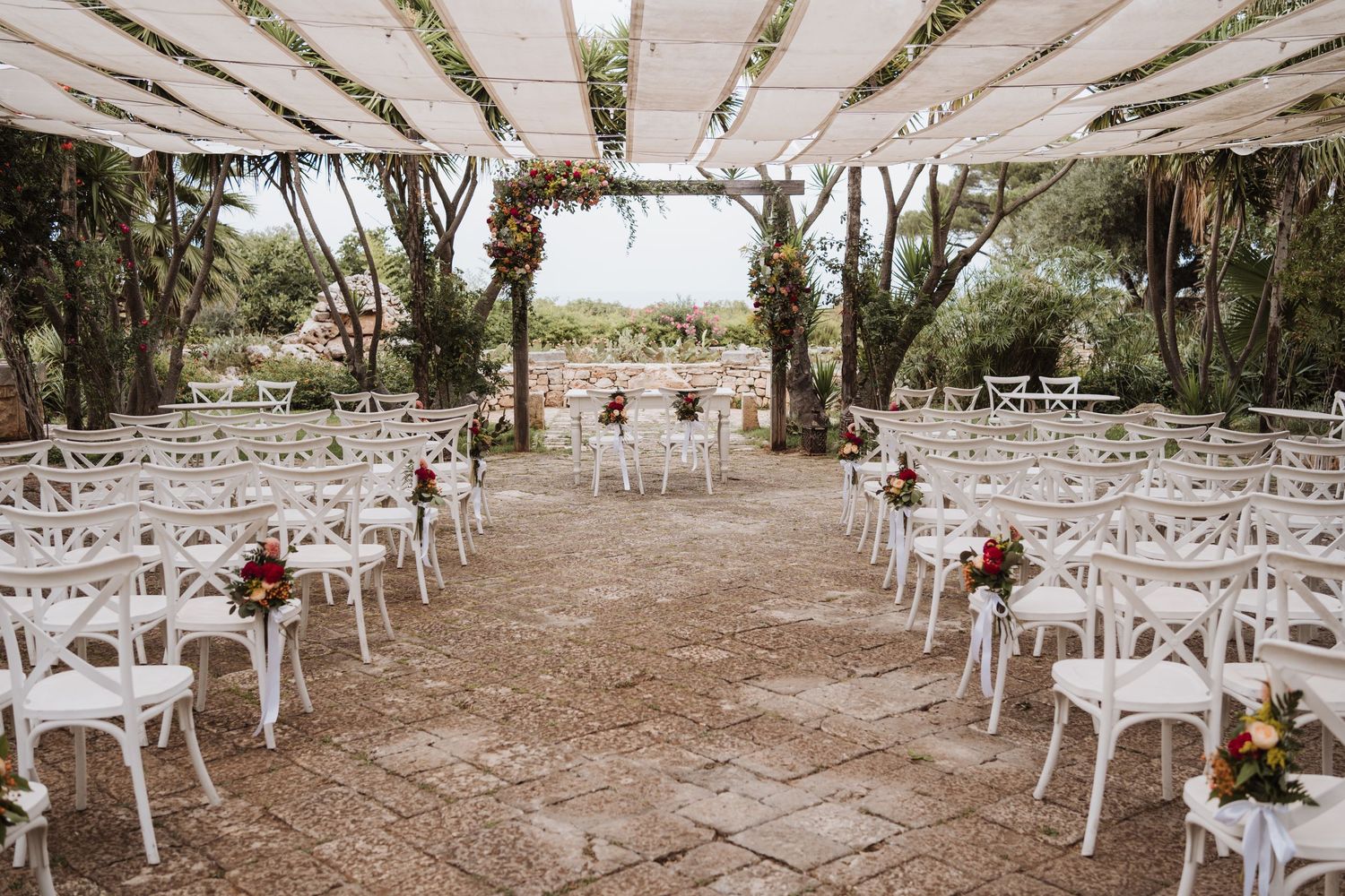 Outdoor wedding ceremony setup with white chairs and floral arch under a draped canopy.