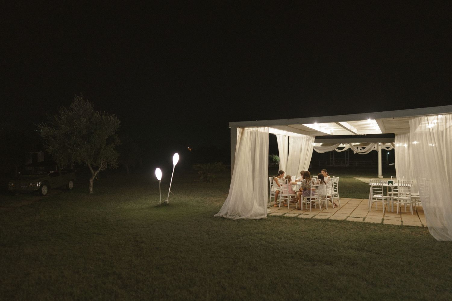 A lit outdoor reception tent glows in the darkness with string lights and guests inside.