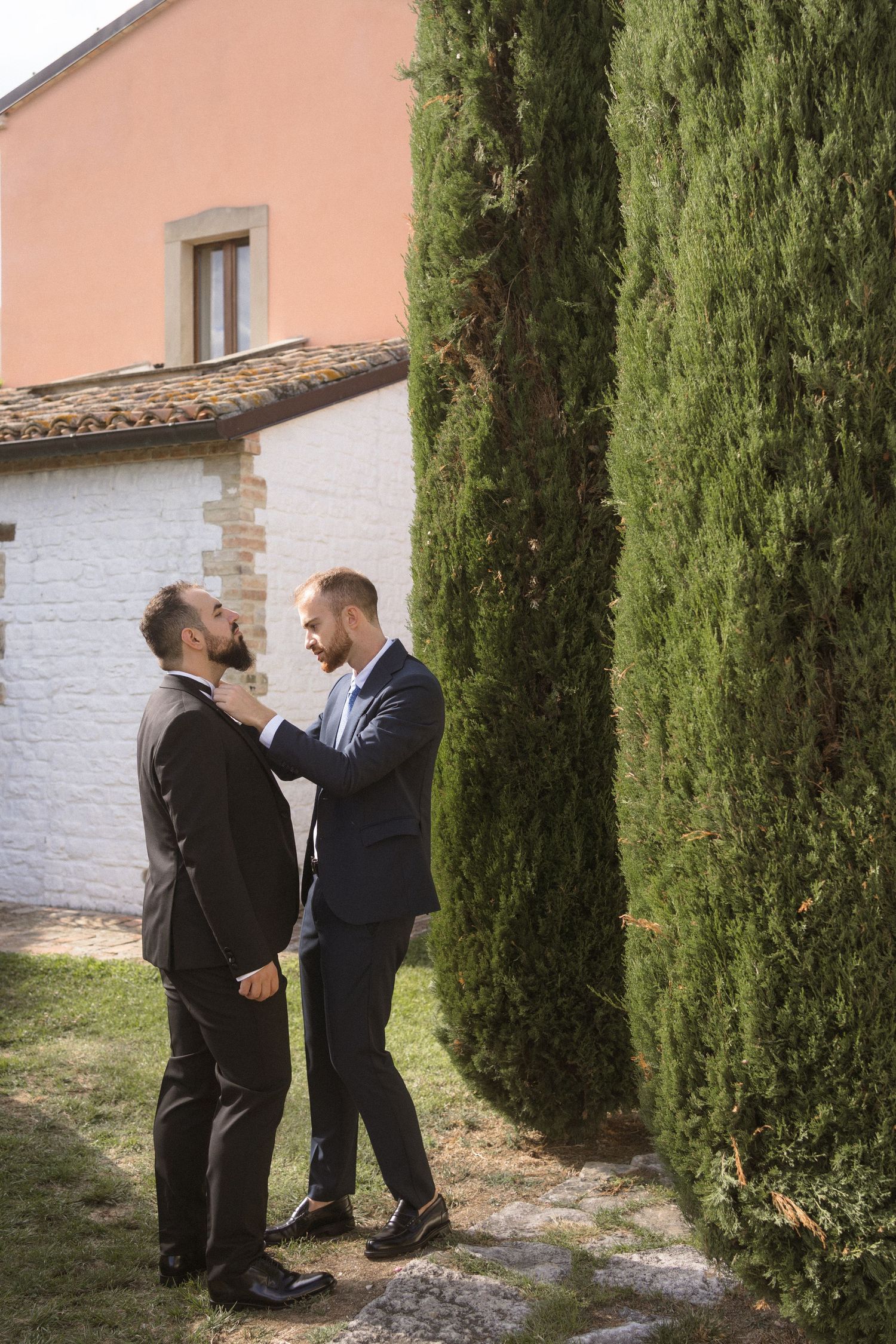 Two men in formal attire having an emotional conversation between cypress trees outside a building.