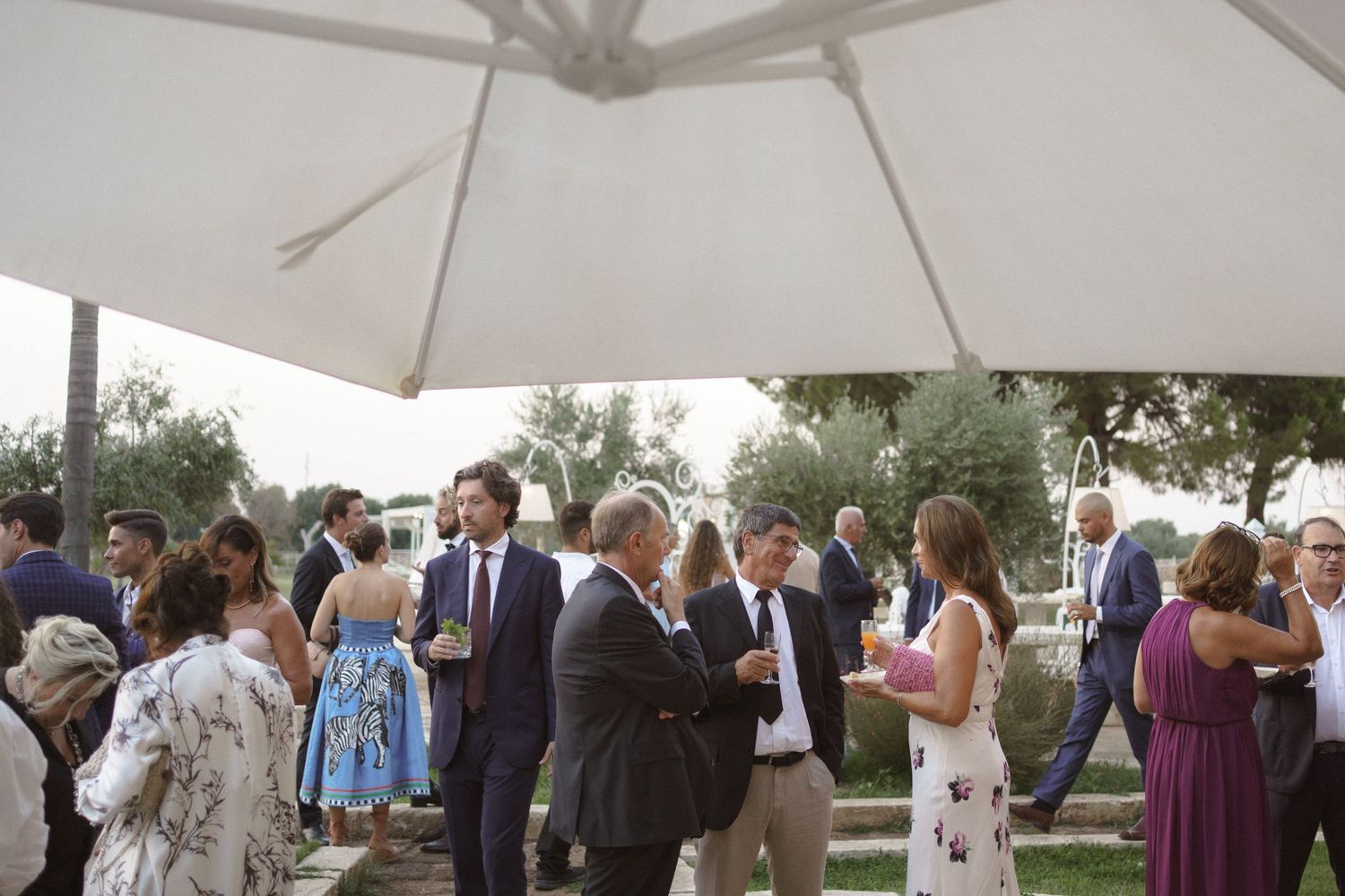 Wedding guests mingling under a white umbrella at an outdoor reception.