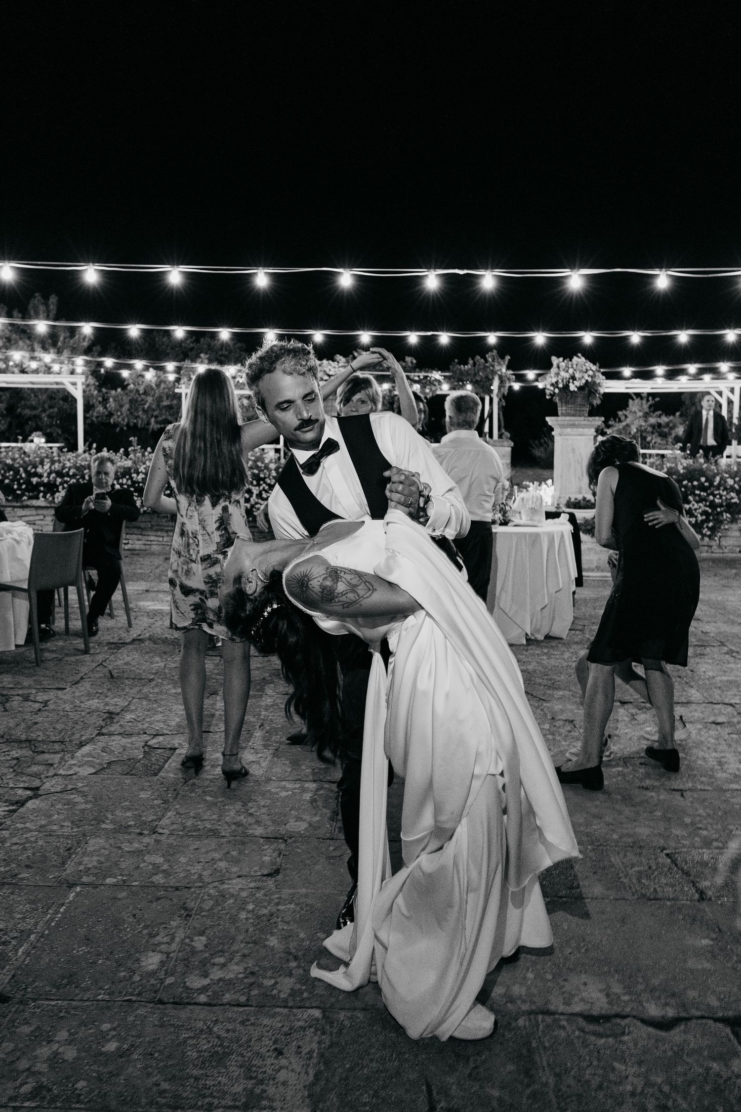 Wedding couple shares a romantic dance outdoors under string lights at an evening reception celebration.
