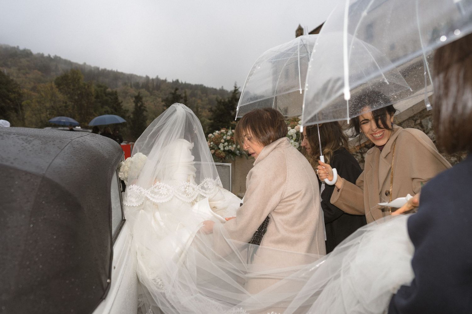 Bride in white wedding dress and veil arriving in the rain with helpers.