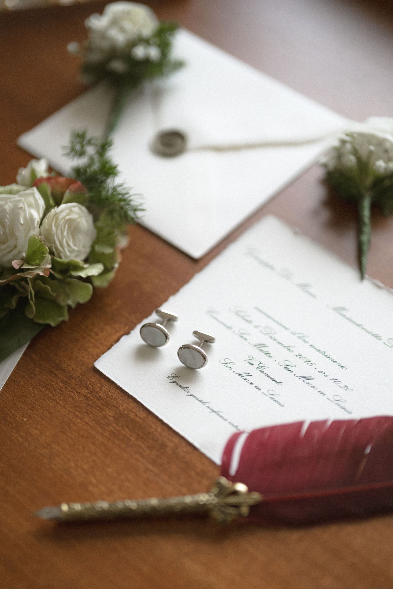 Wedding stationery flat lay with white floral boutonnieres, cufflinks, wax seal envelope, and red quill pen on wooden table.