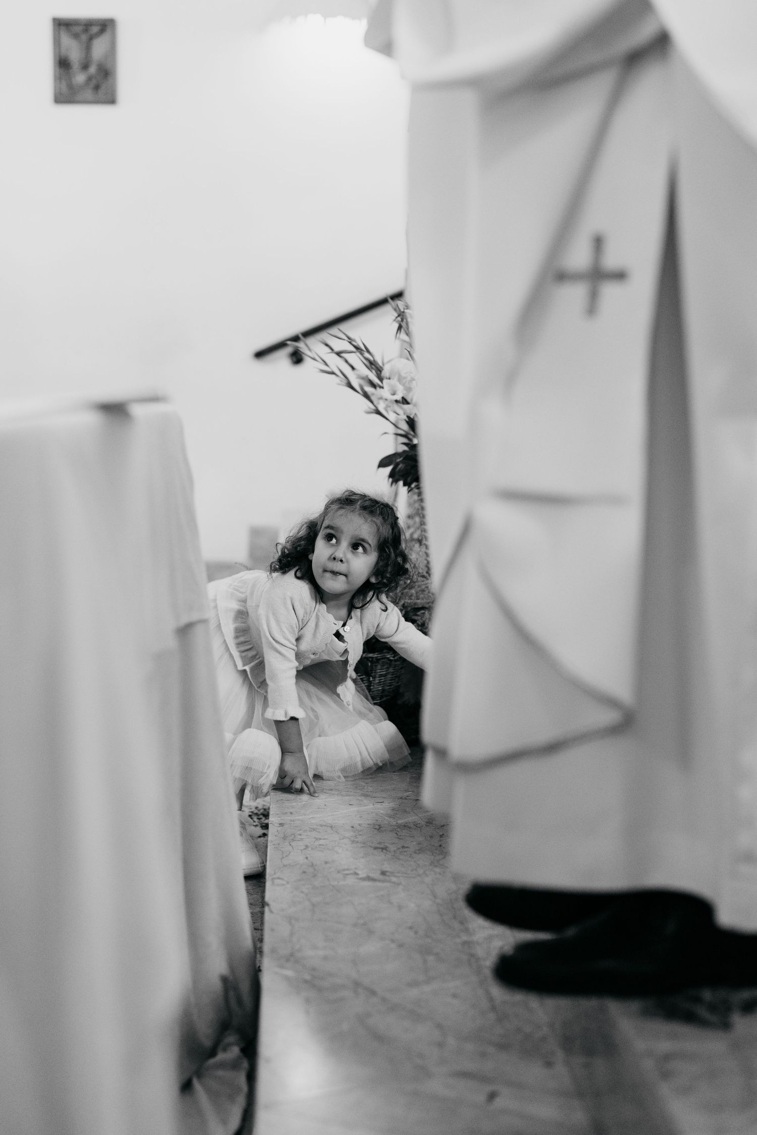 Black and white photo of a young girl in white dress sitting on church floor, looking up among robed clergy figures.