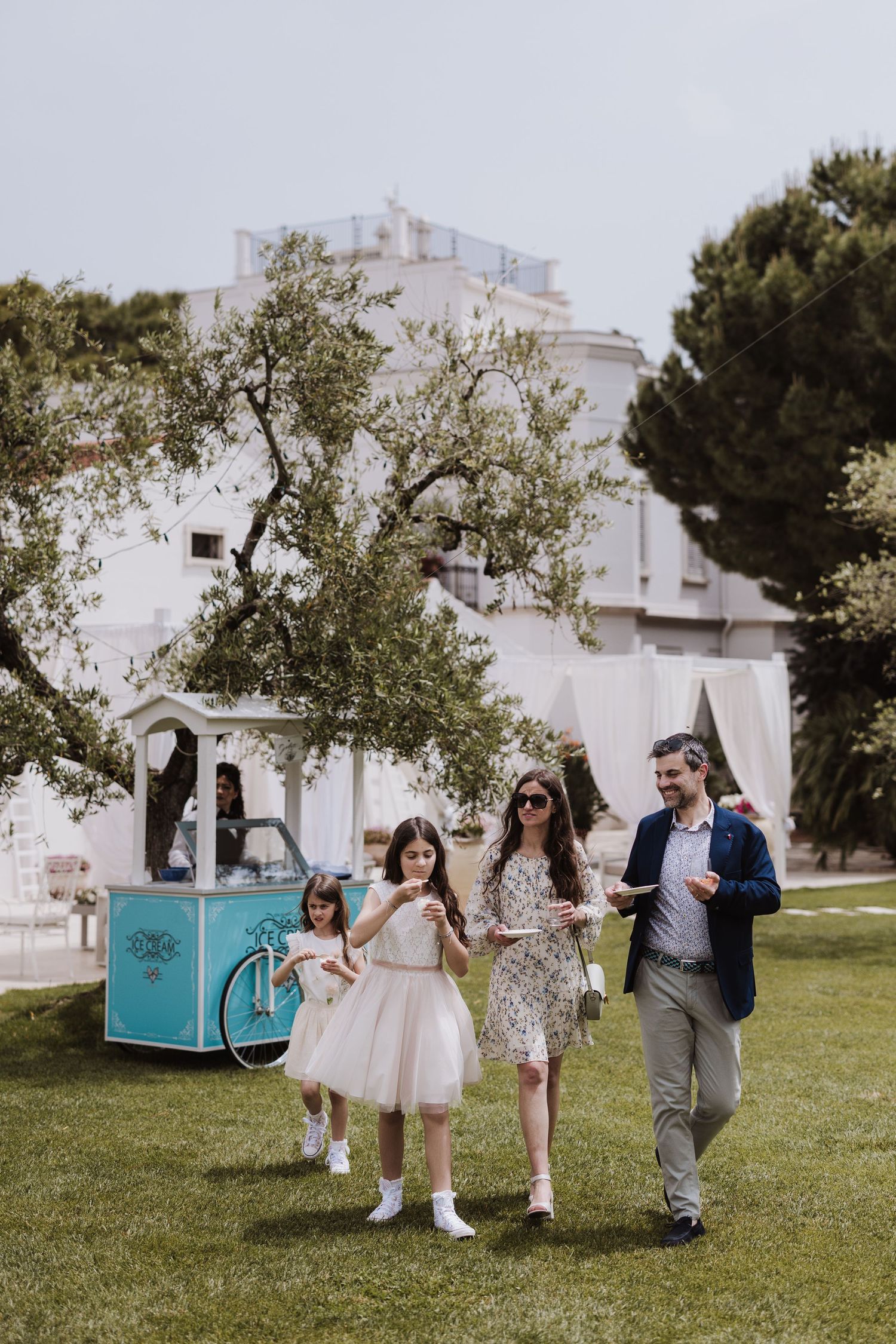 Family walking on lawn with vintage blue ice cream cart and white building in background.