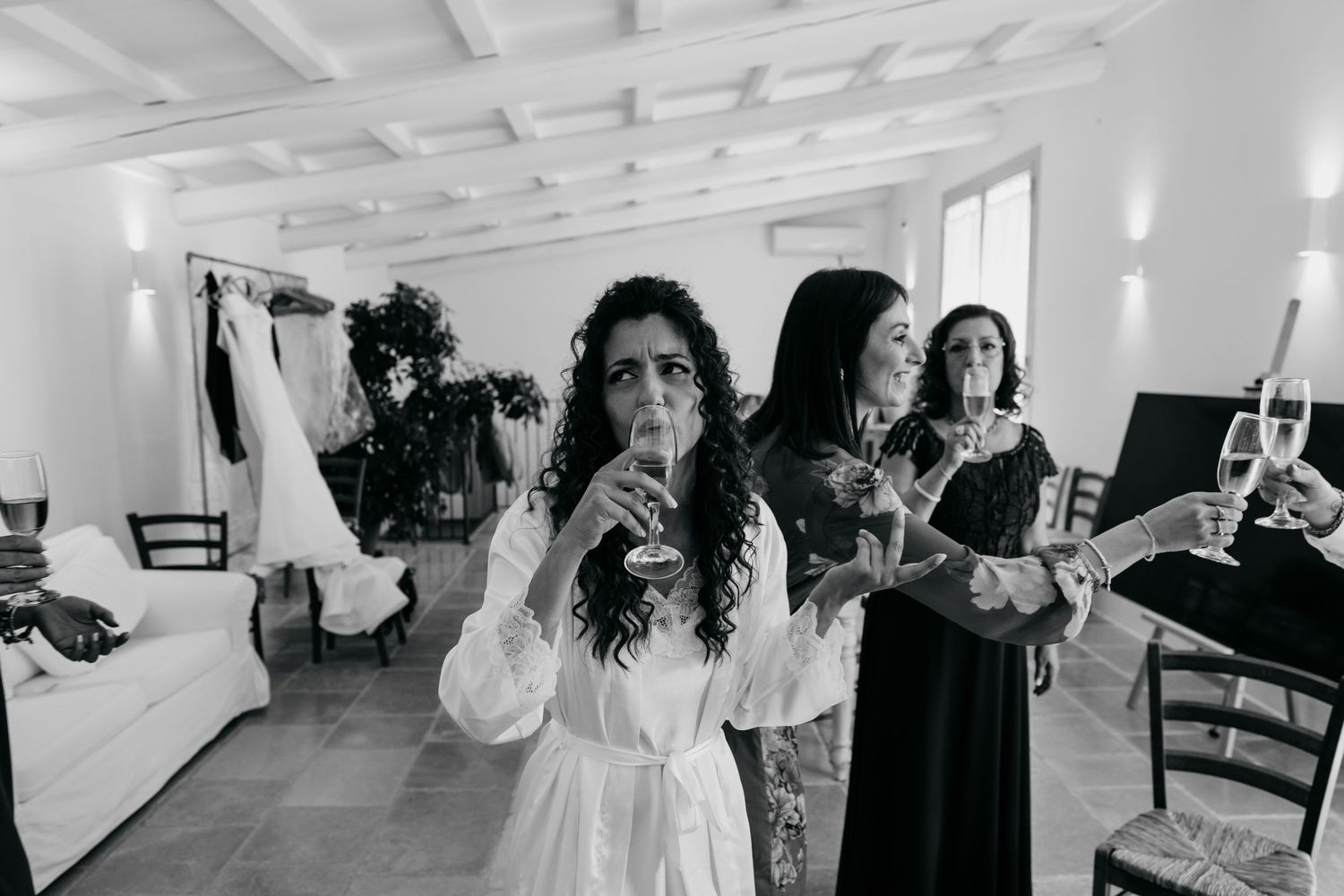 Wedding preparation scene with women in white and black dresses sharing drinks indoors.