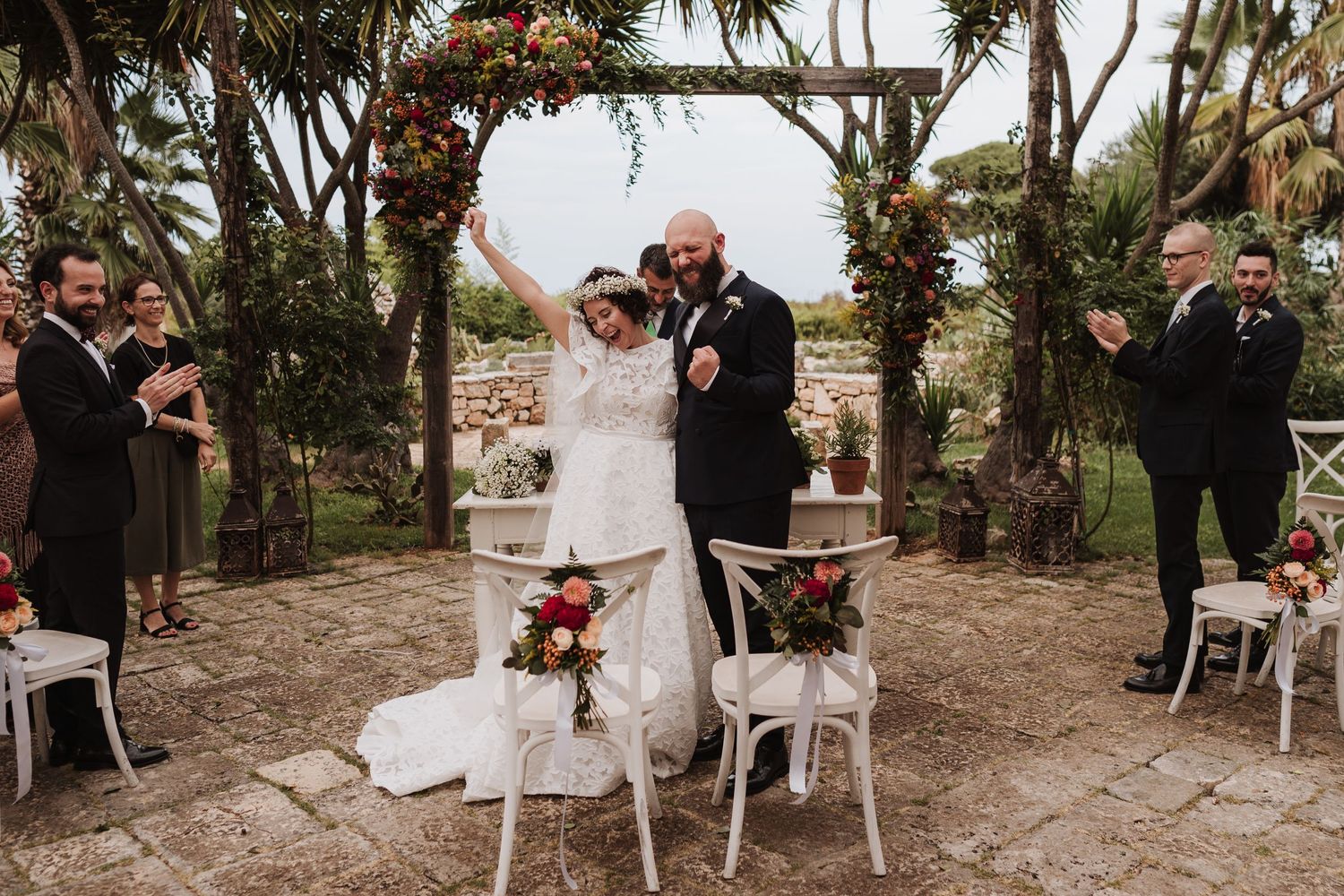 Wedding celebration under wooden arch decorated with flowers as couple raises arms in joy.