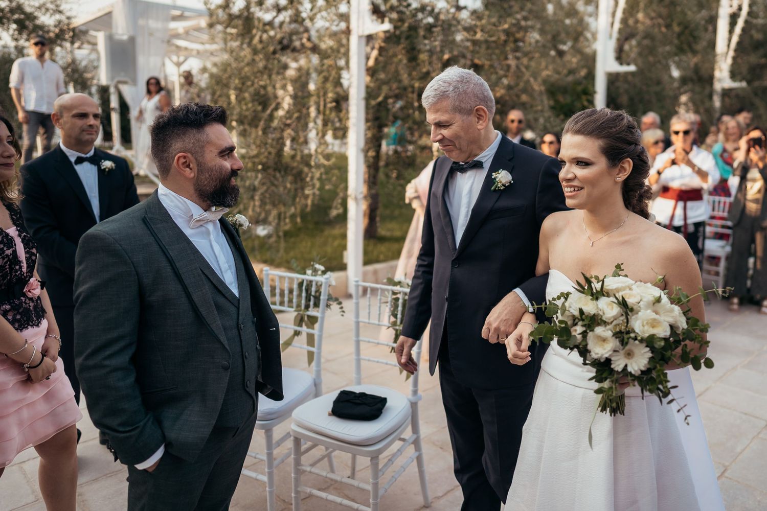 A wedding ceremony takes place outdoors as guests watch the bride walk down the aisle.