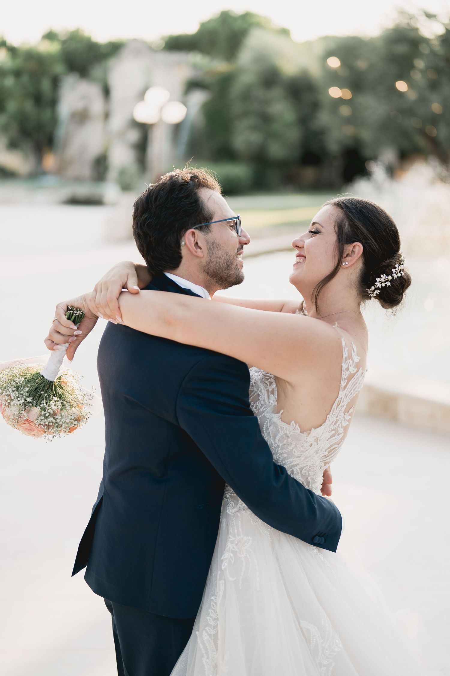 A joyful wedding couple embraces and laughs together in an outdoor setting during sunset.