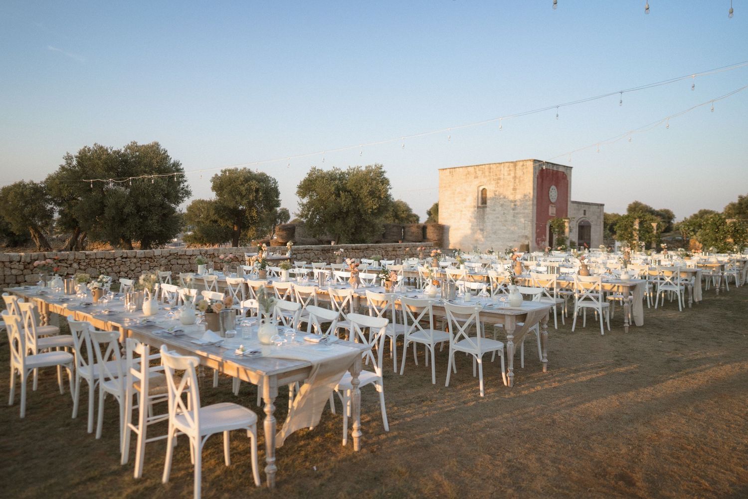 Outdoor wedding reception setup with white chairs and tables under open sky at dusk.