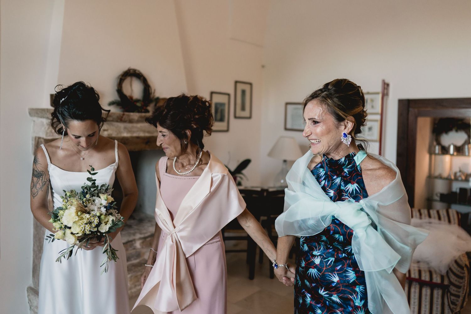 Three women dressed in wedding attire share a candid moment in a brightly lit room before the ceremony.