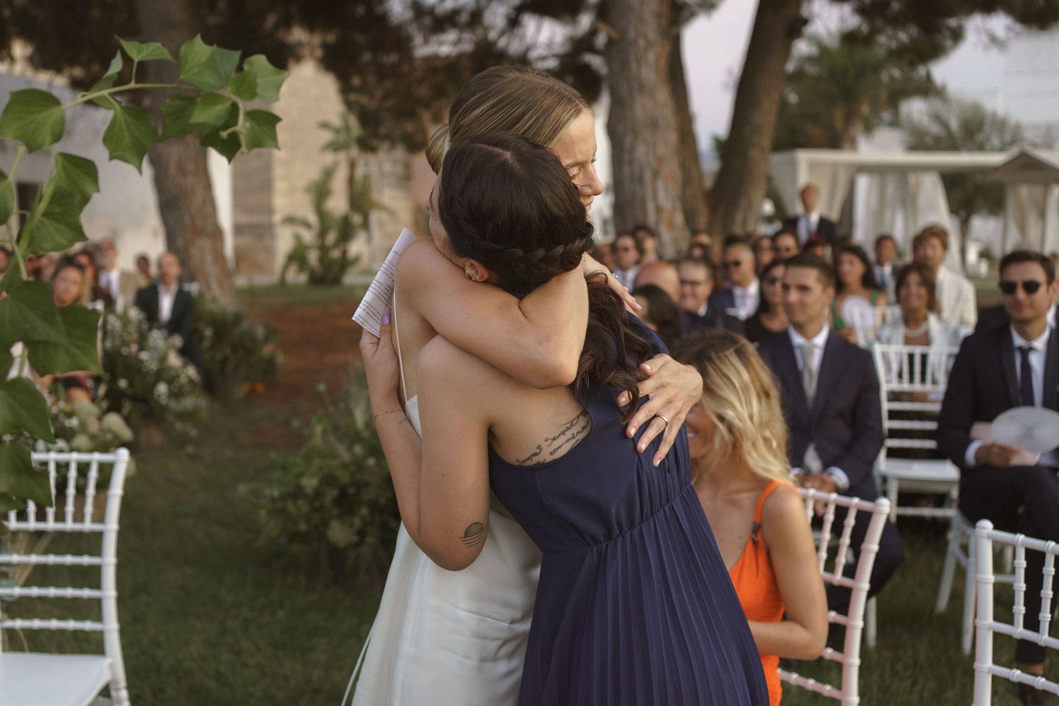 A bridesmaid in navy blue hugs someone in a white gown during an outdoor wedding ceremony with white chairs.