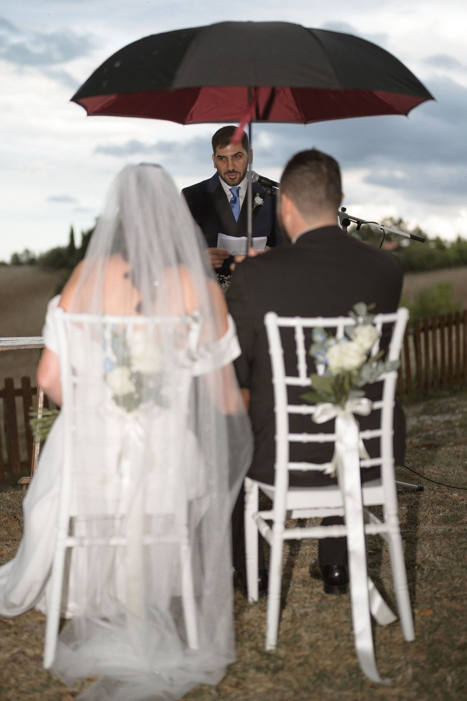 Wedding ceremony chairs and decorations under umbrellas on a cloudy day in an outdoor setting.