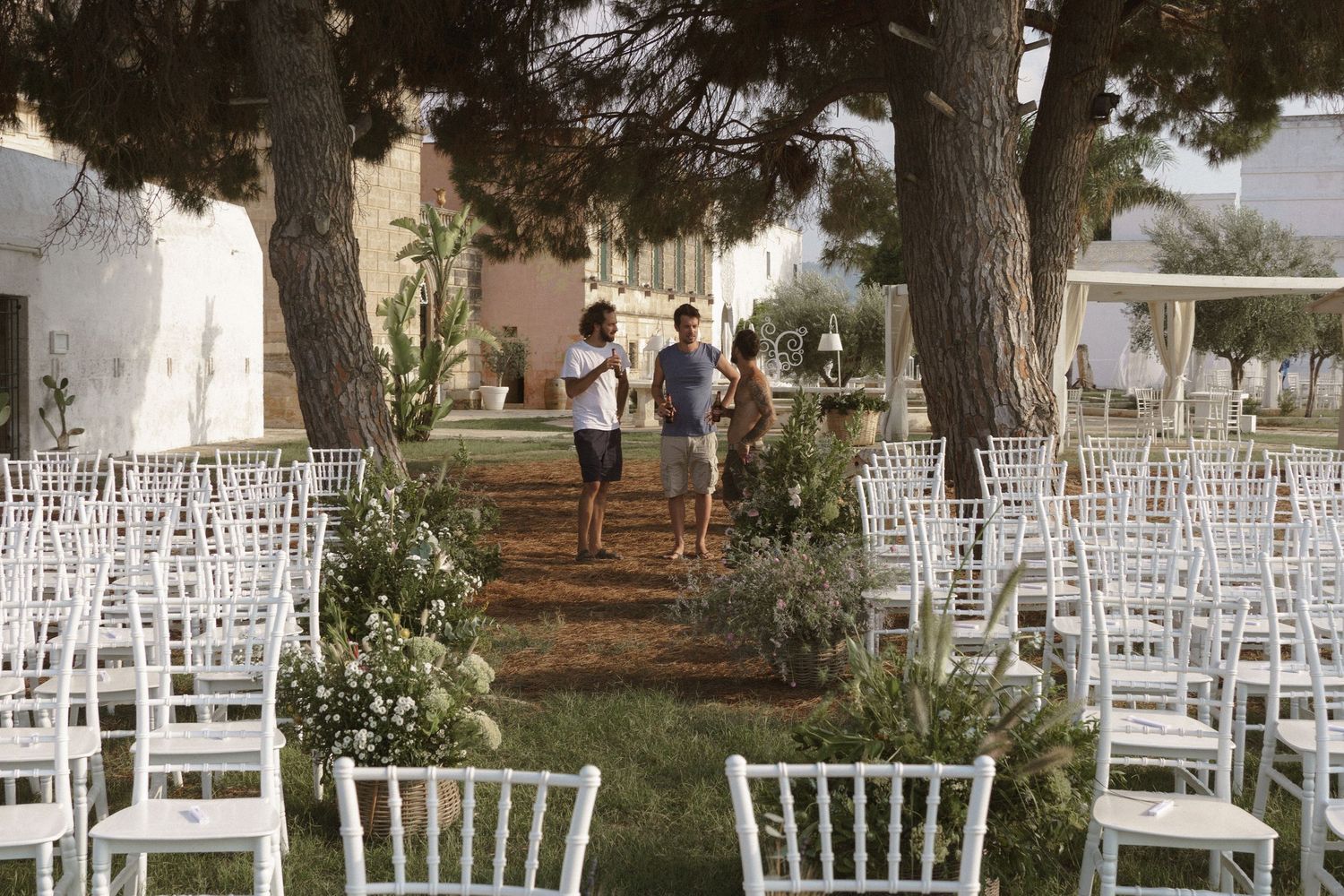 White chiavari chairs arranged outdoors for a garden wedding ceremony with pine trees and greenery lining the aisle.