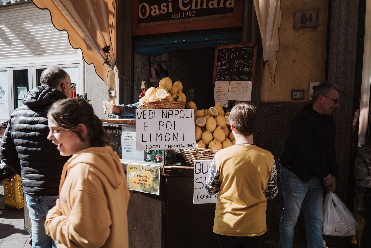 People queue at a small bread shop in Naples with fresh loaves and handwritten signs displayed at the storefront entrance.