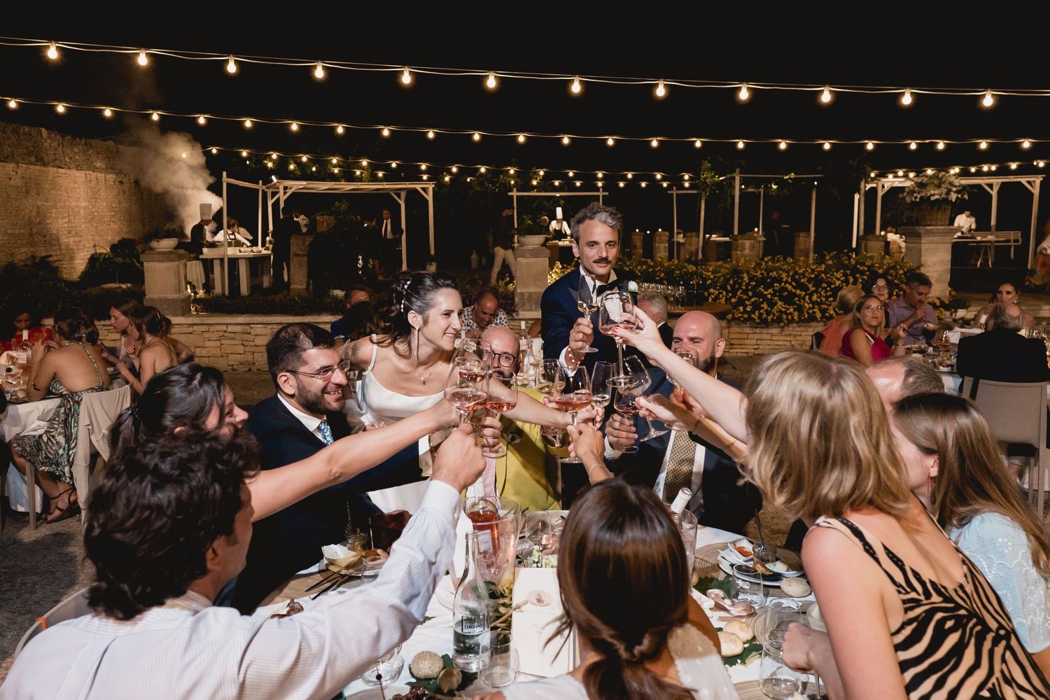 Wedding guests raise their glasses in a toast during outdoor reception under twinkling string lights.