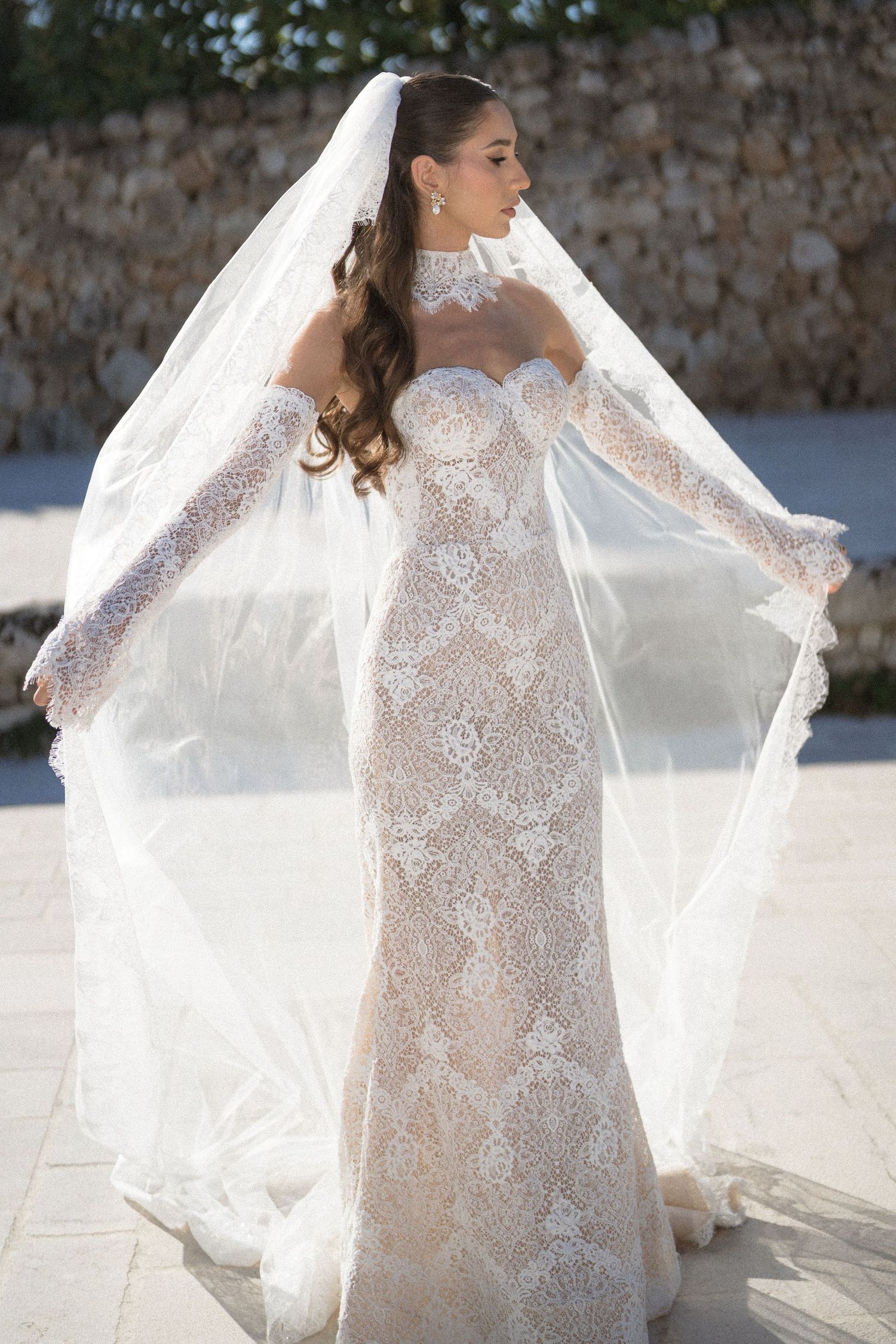 Bride in a fitted lace wedding dress lifts her sheer veil outdoors beside a stone wall, wearing long lace gloves.