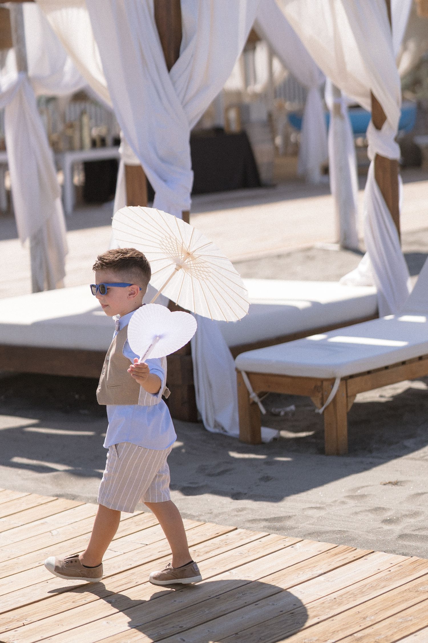 Young ring bearer in sunglasses walking with parasol at beach wedding.