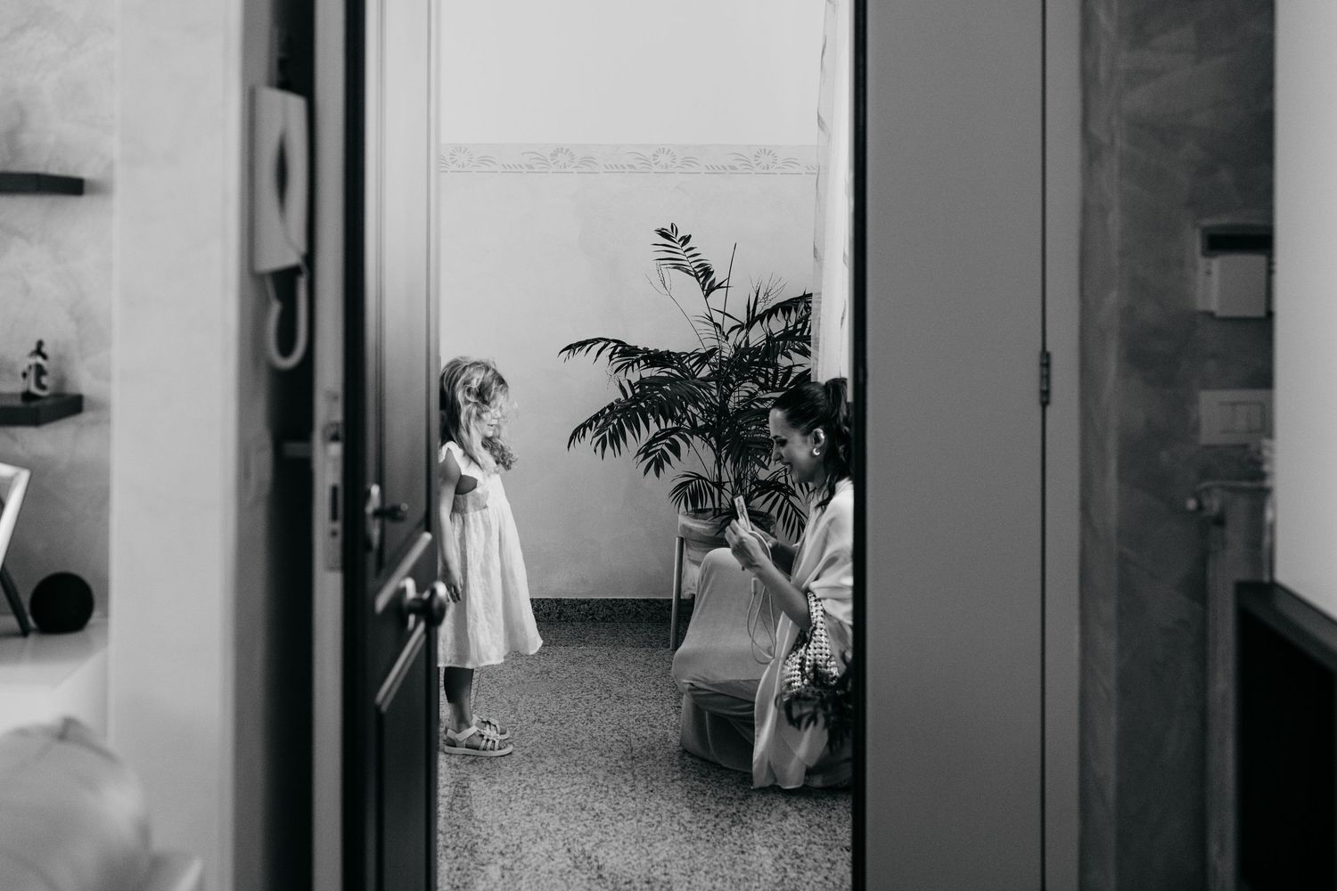 Black and white photo of a woman kneeling to adjust a little girls white flower girl dress before a wedding.