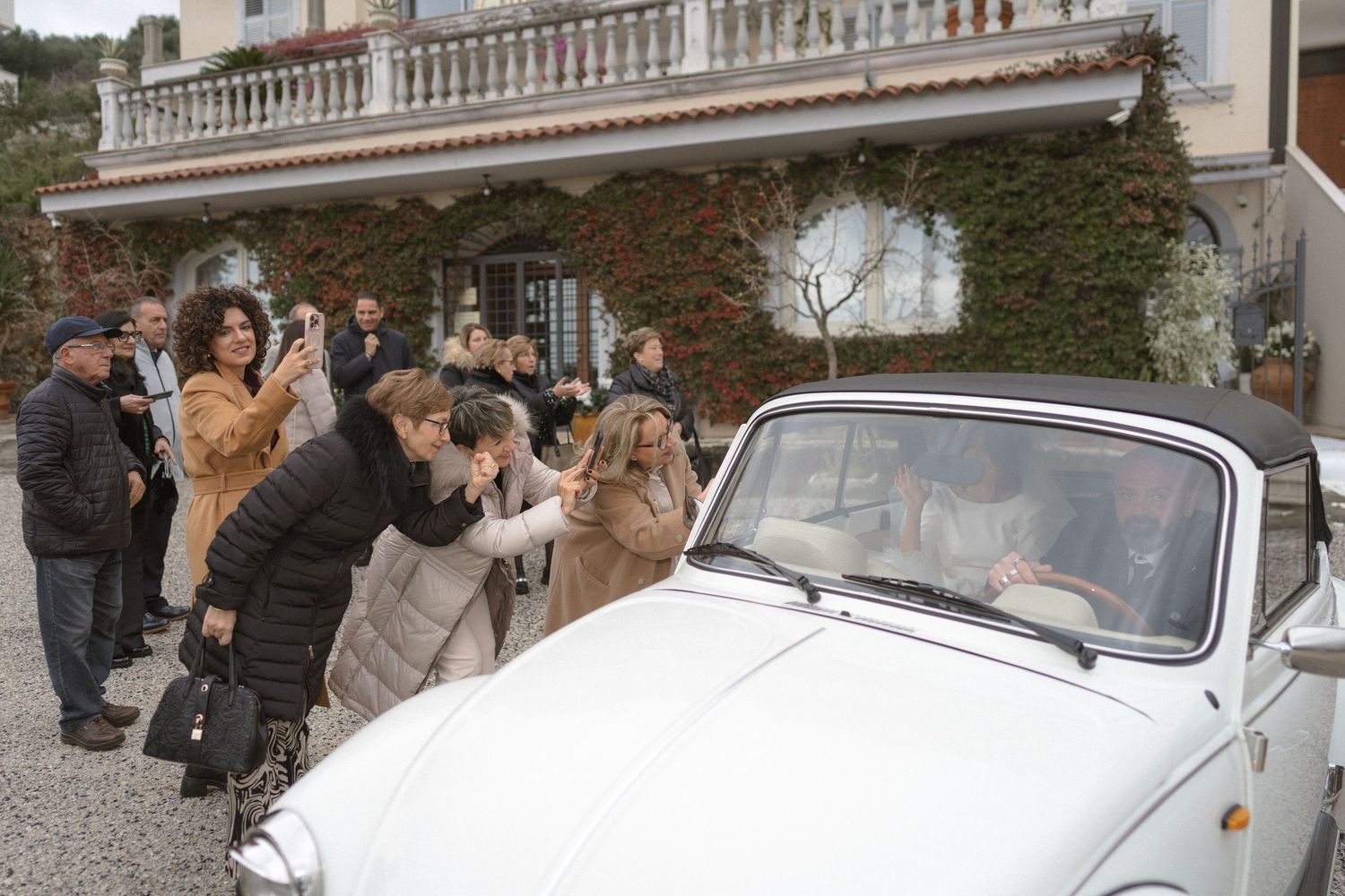 Guests photograph a bride and groom in a classic white convertible car outside an elegant villa.