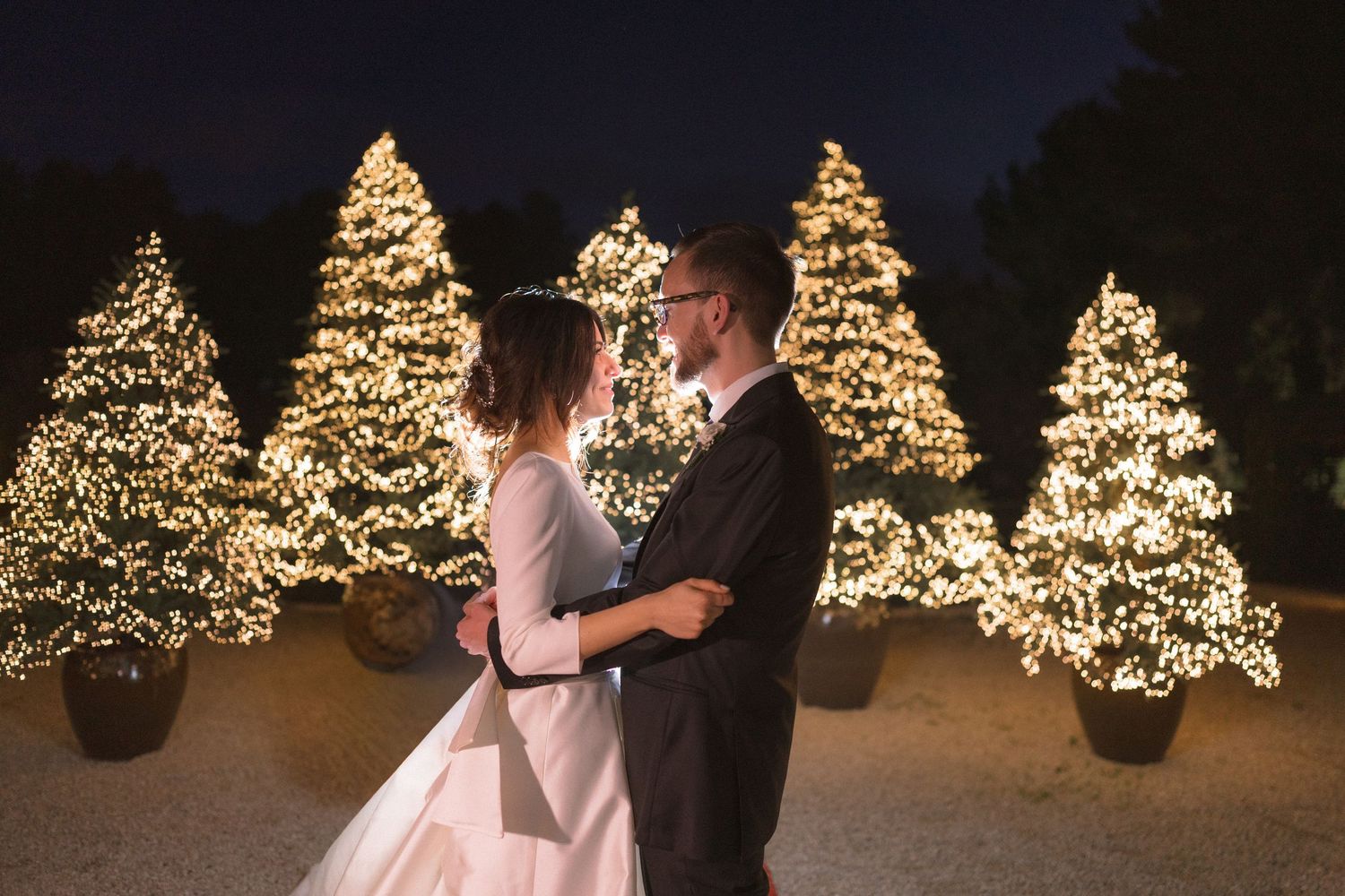 Romantic couple embracing before illuminated Christmas trees with warm fairy lights at night wedding.