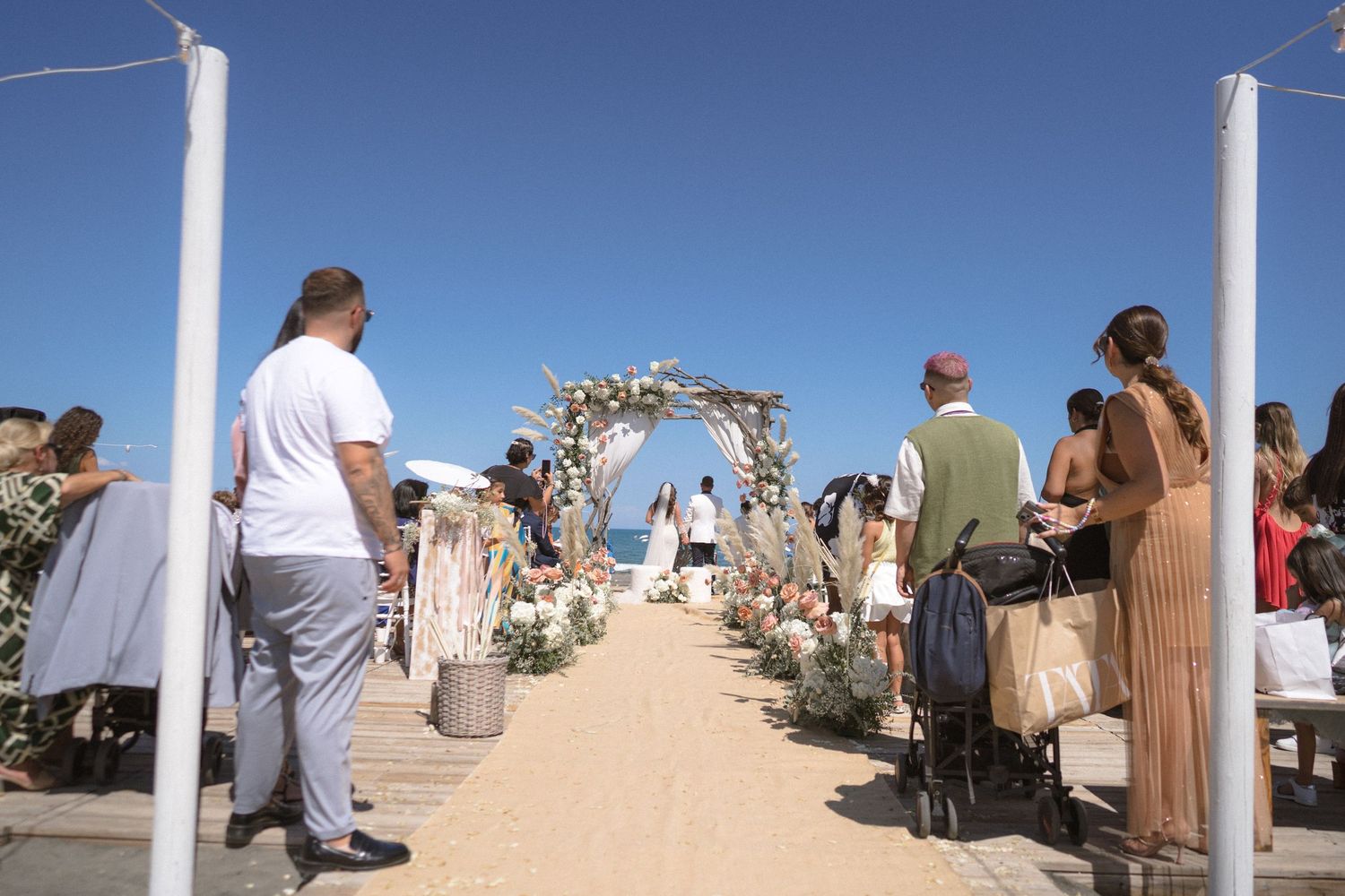 White wedding arch and chairs set up on sandy beach with blue sky backdrop.