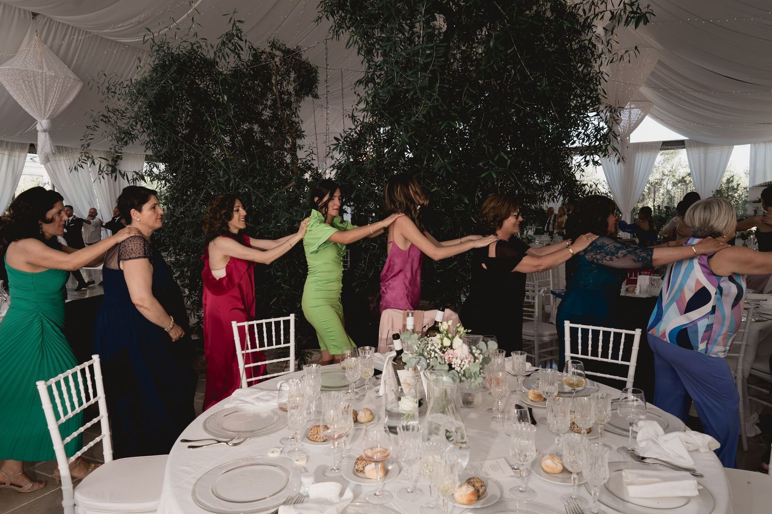 Wedding guests in colorful dresses form a dance circle at an elegant reception.