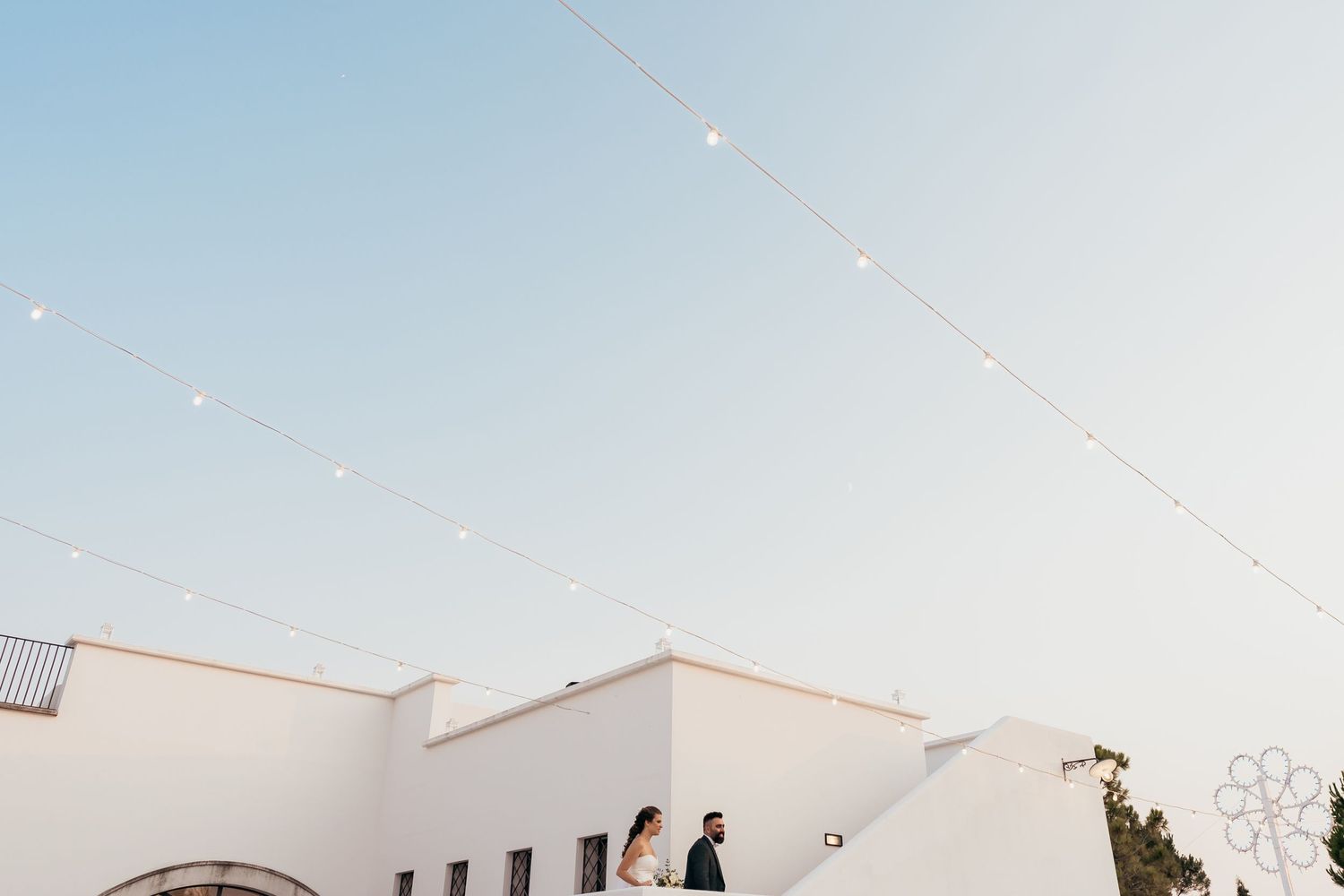 String lights hang above a white Mediterranean-style building at dusk.