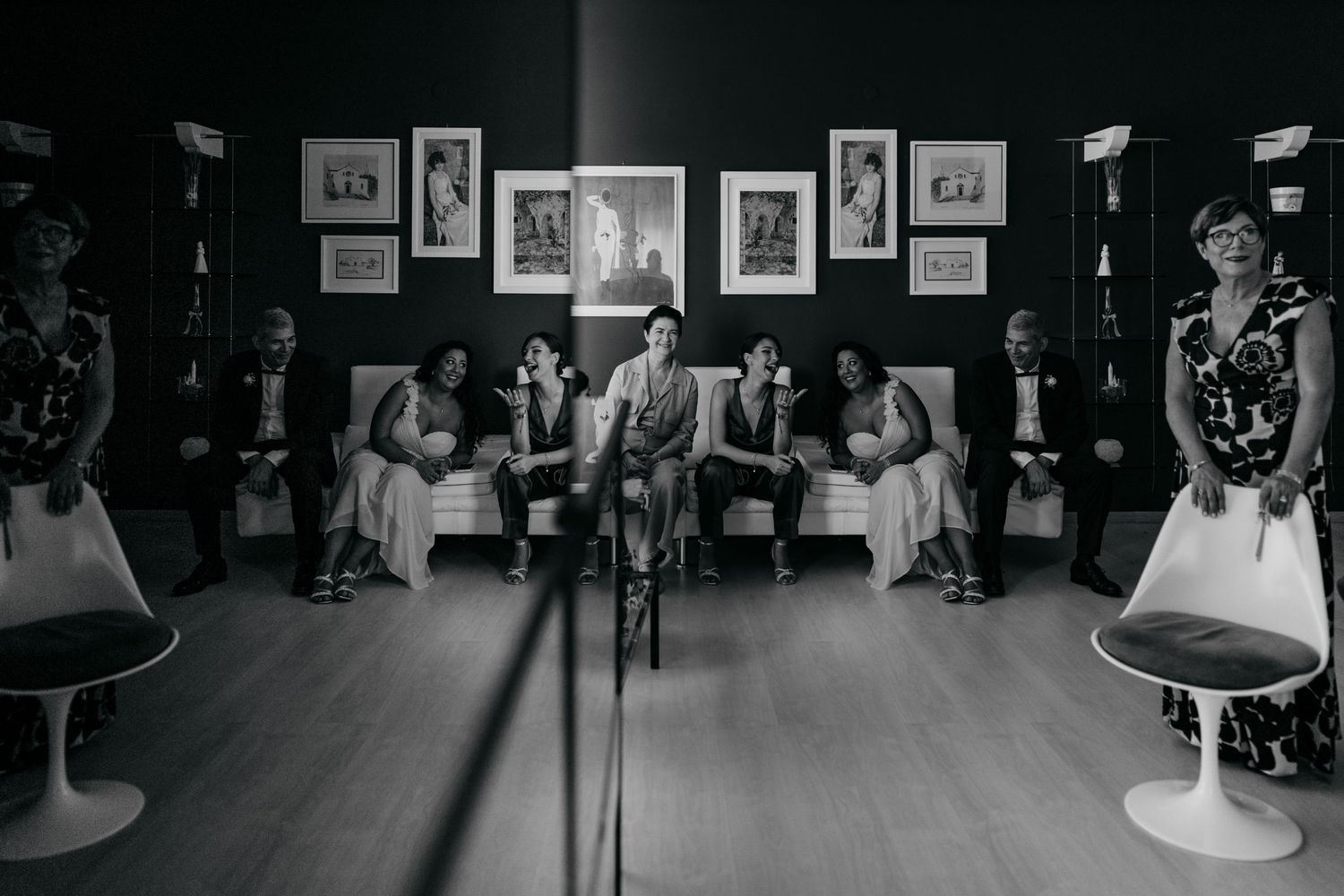 Black and white photo of a wedding party seated in a room with framed photos on the wall.