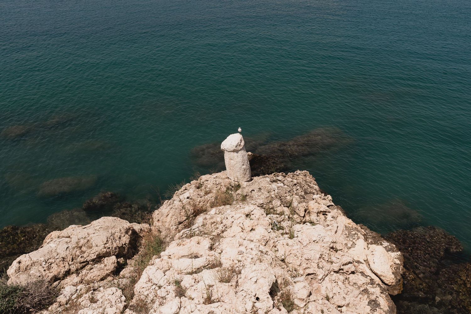 Lone figure sits on rocky cliff overlooking deep turquoise Mediterranean waters.