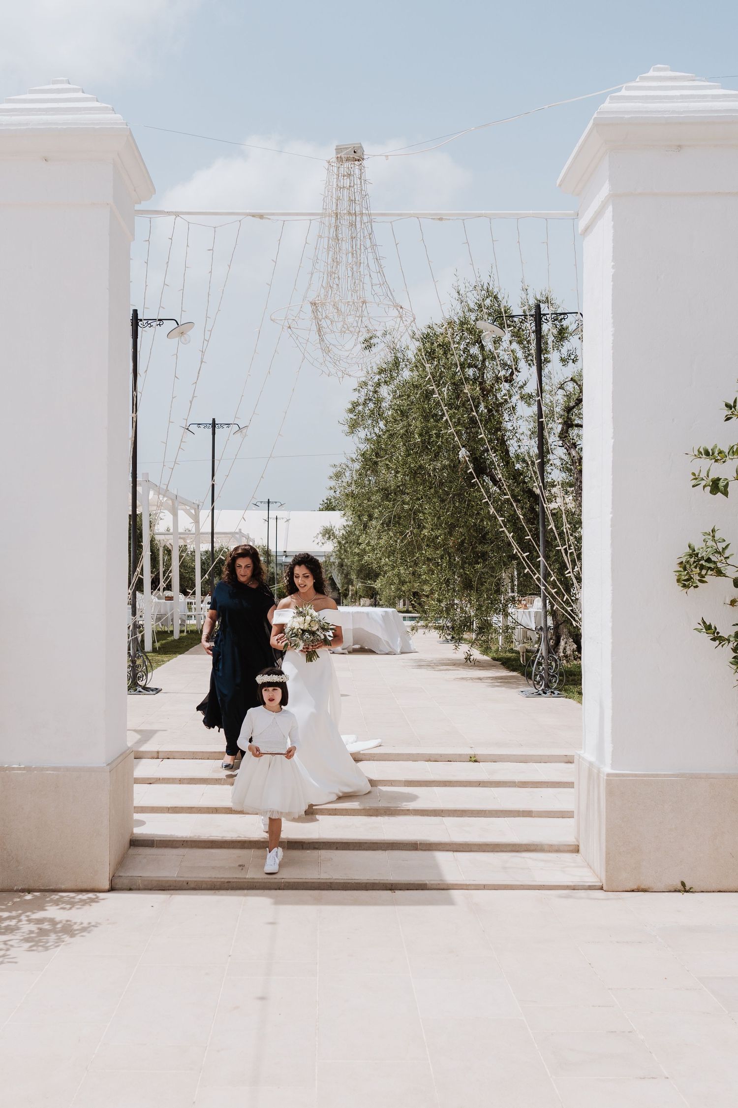 Wedding party descends white steps under an archway with power lines visible.