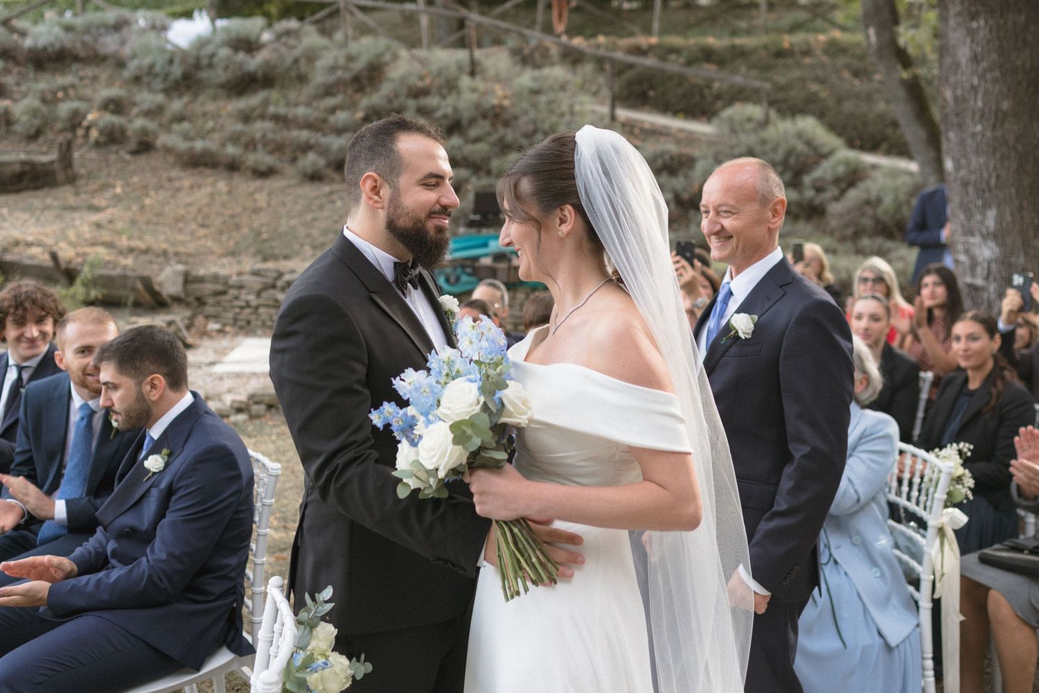 A joyful outdoor wedding ceremony takes place in a scenic wooded setting with guests seated on white chairs.