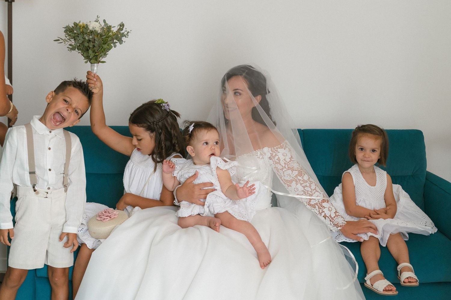 Children in white clothing sit together on a teal couch during a special occasion.