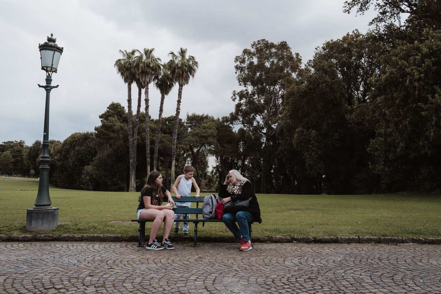People relaxing on a bench in a park surrounded by palm trees and a vintage street lamp.
