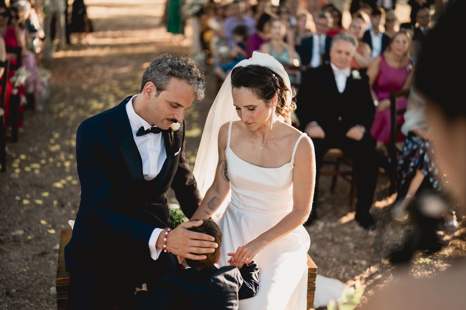 An elegant outdoor wedding ceremony shows a couple sitting together on autumn day with guests in background.