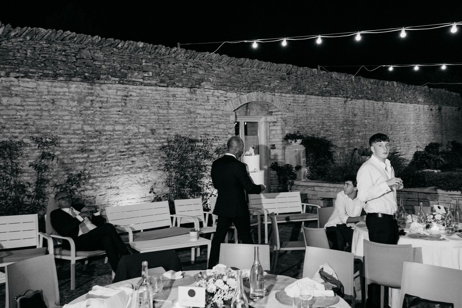 Black and white photo of an outdoor reception venue with string lights and place settings on tables near a brick wall.