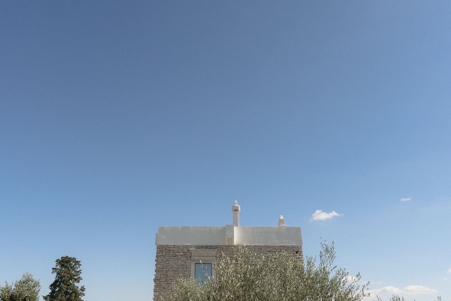Rustic stone building with chimney against clear blue sky surrounded by olive trees.