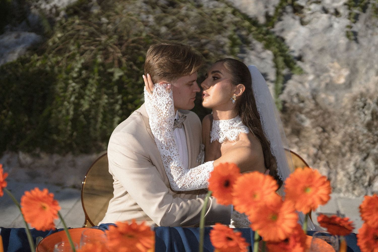 Groom in a cream suit embraces a veiled bride among bright orange gerbera daisies in a rocky natural setting.