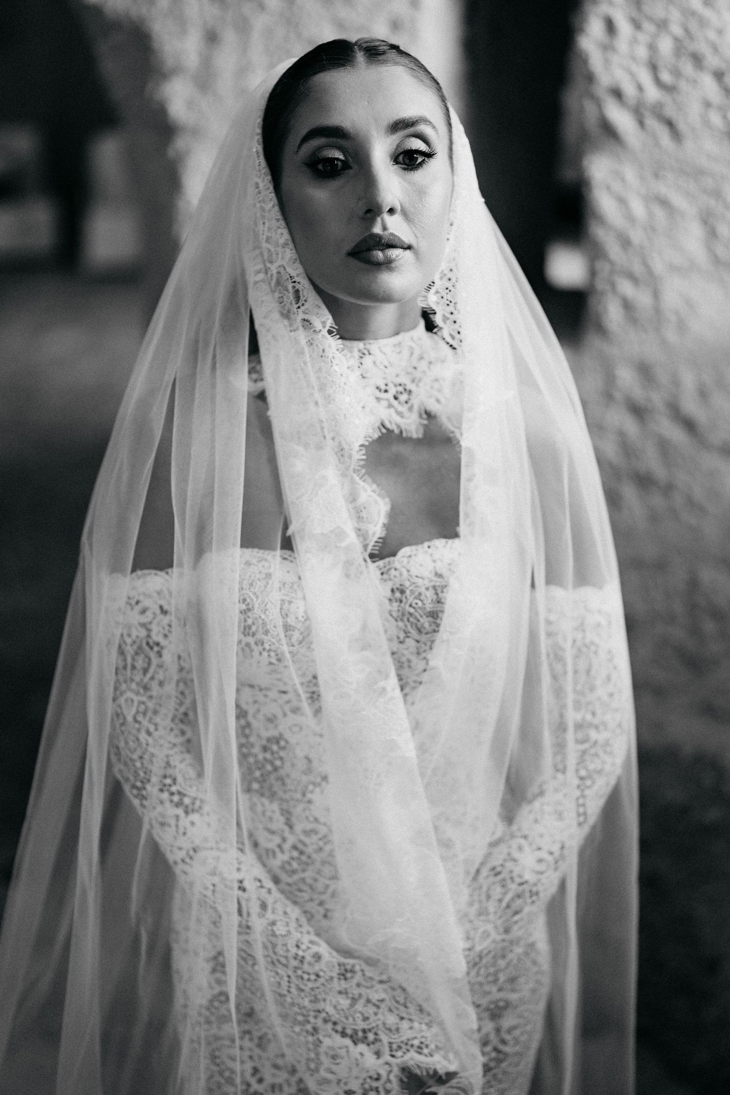 Black and white portrait of a bride wearing a sheer lace-trimmed veil and an off-shoulder lace wedding dress.