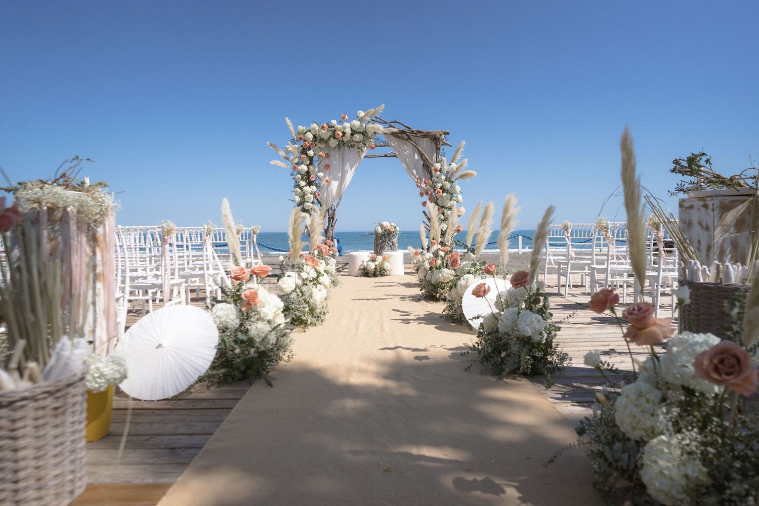 Romantic beach wedding ceremony setup with floral arch and white chairs on sandy path.