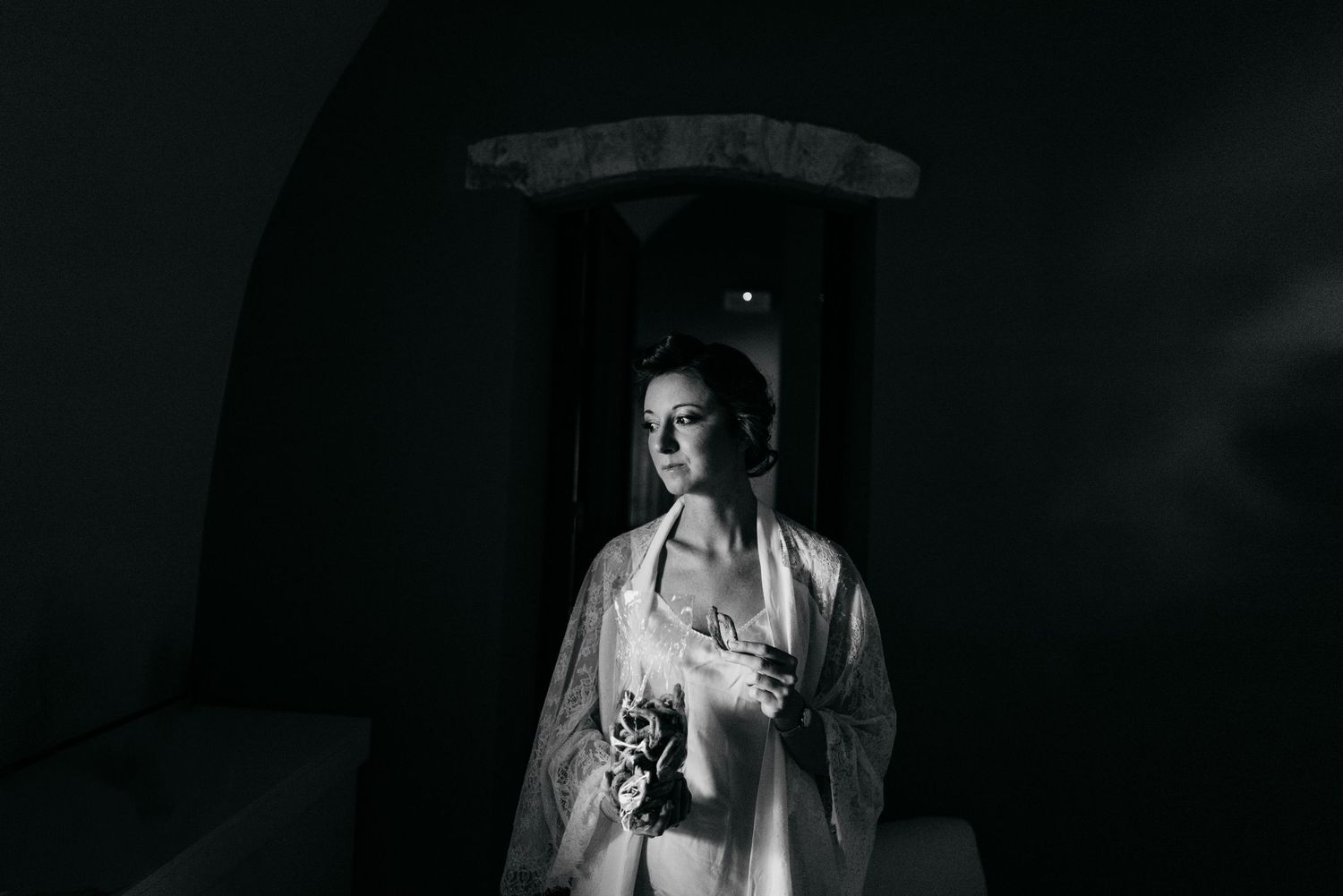 Black and white bridal portrait of elegant woman in silk robe holding bouquet, standing in dramatic arched doorway.