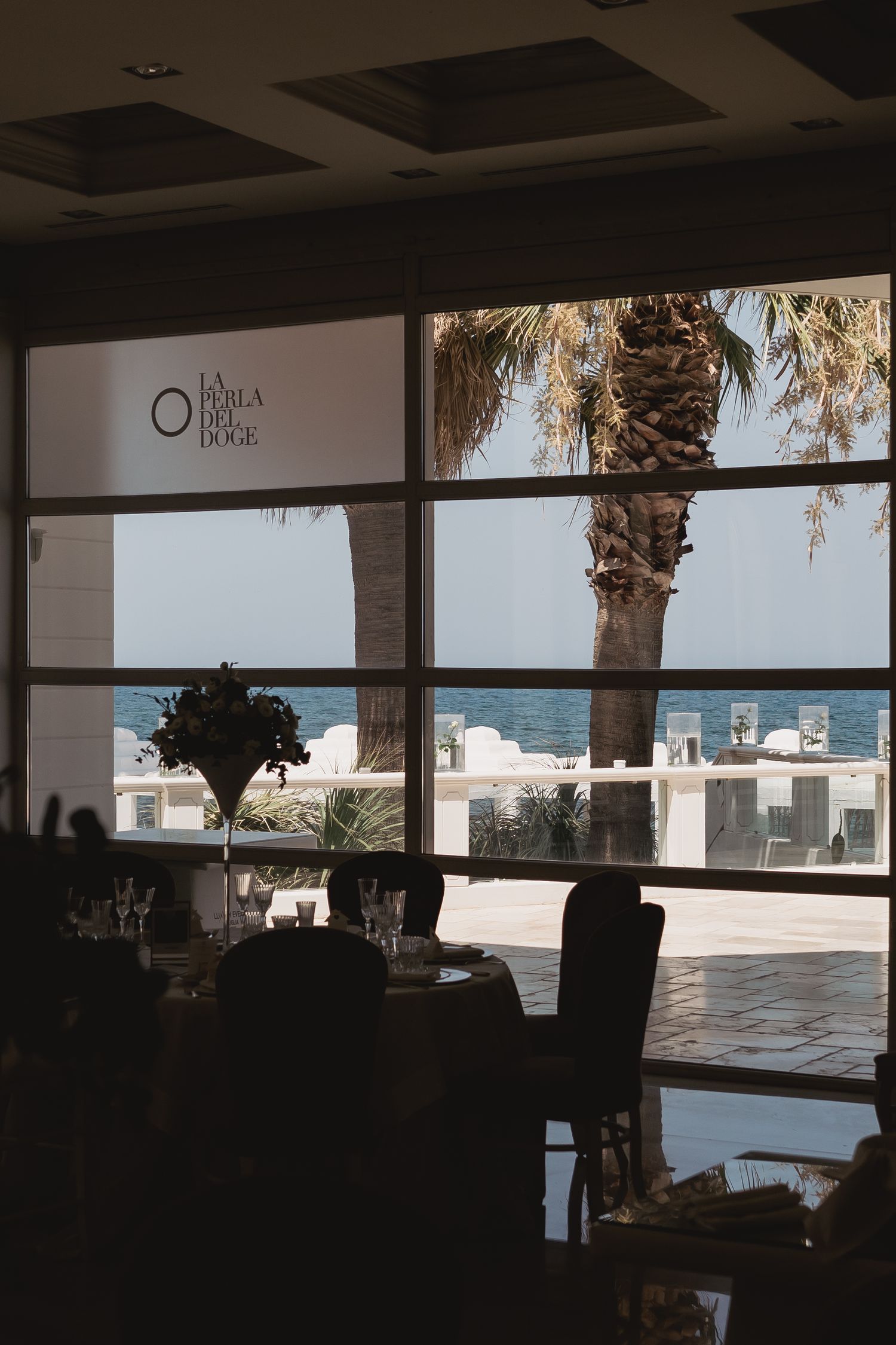 View through a restaurant window overlooking a beachfront with palm trees and white buildings against the ocean.