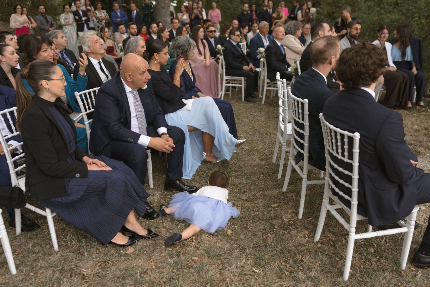Wedding guests seated in white chairs at an outdoor ceremony.