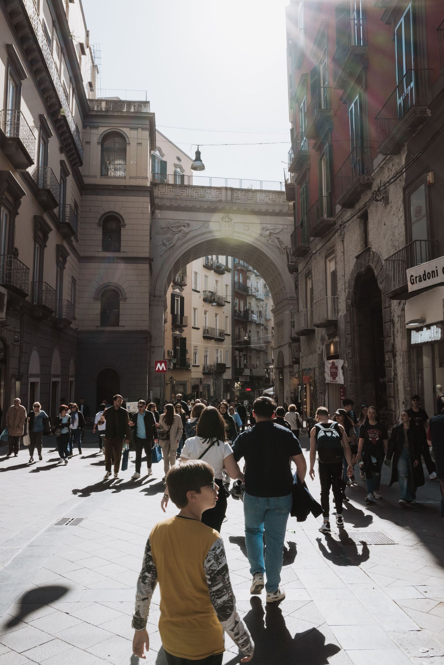 A busy pedestrian street in a European city with historic architecture and an ornate stone archway.