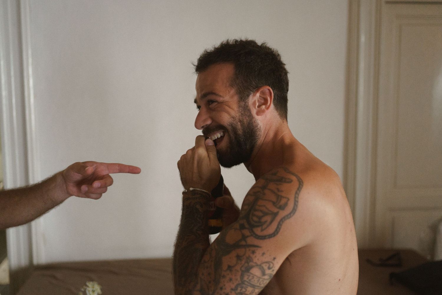 A shirtless person with tattoos laughs while someone points at them in a light-filled room.
