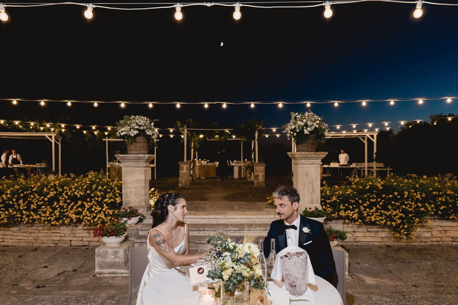 A couple sits at an outdoor reception table under string lights with stone pillars and yellow flowers in the background.