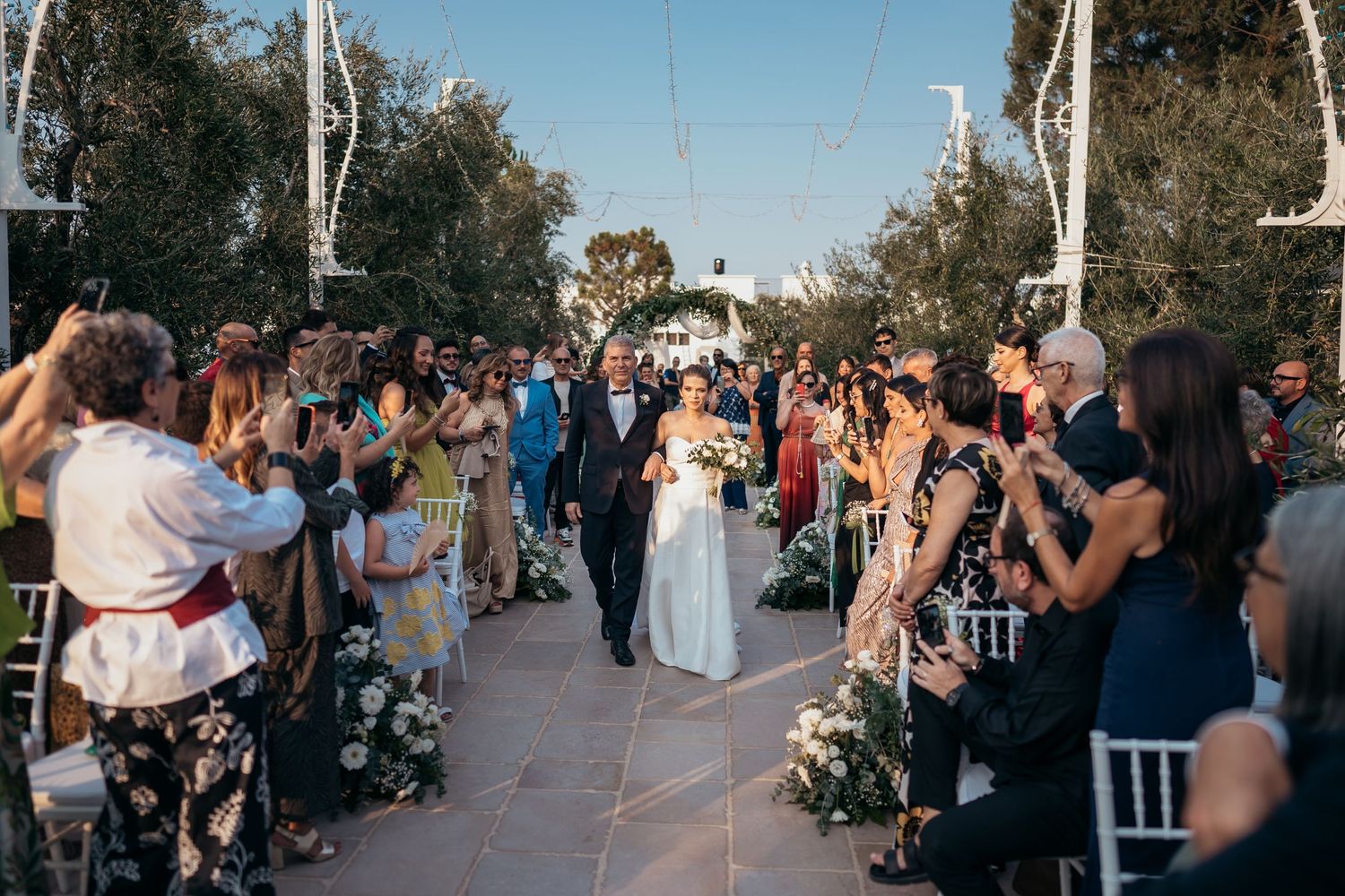 Wedding ceremony aisle lined with guests and white decorations under clear blue sky.