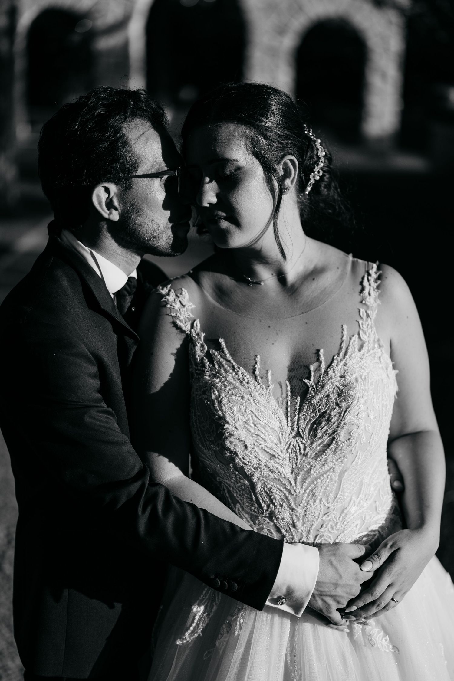A romantic black and white portrait of a couple in wedding attire posed against arched architecture.